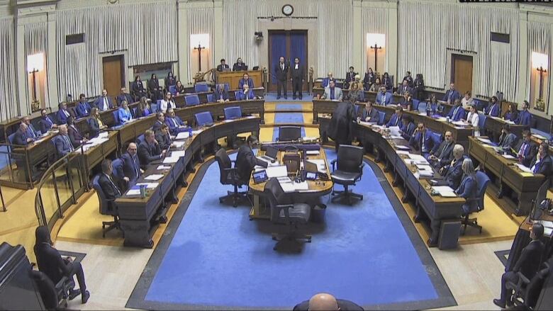 Several people sit in a blue-carpeted legislative chamber.