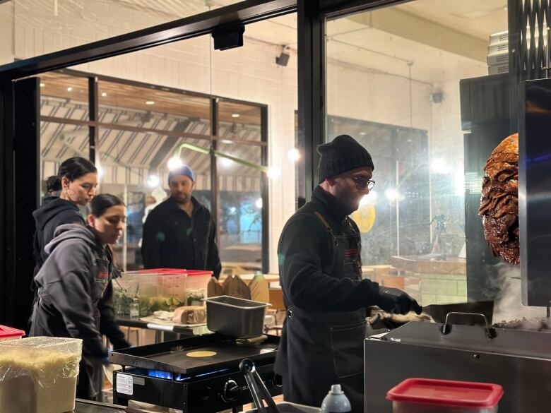 Workers prepare food at a vertical spit while a man looks on.