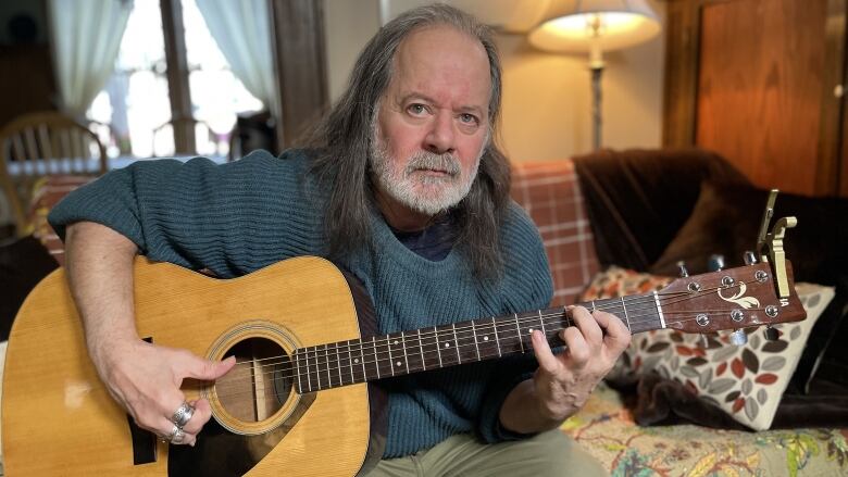 A portrait of a man, holding a guitar with long hair and a sweater, sitting on a couch