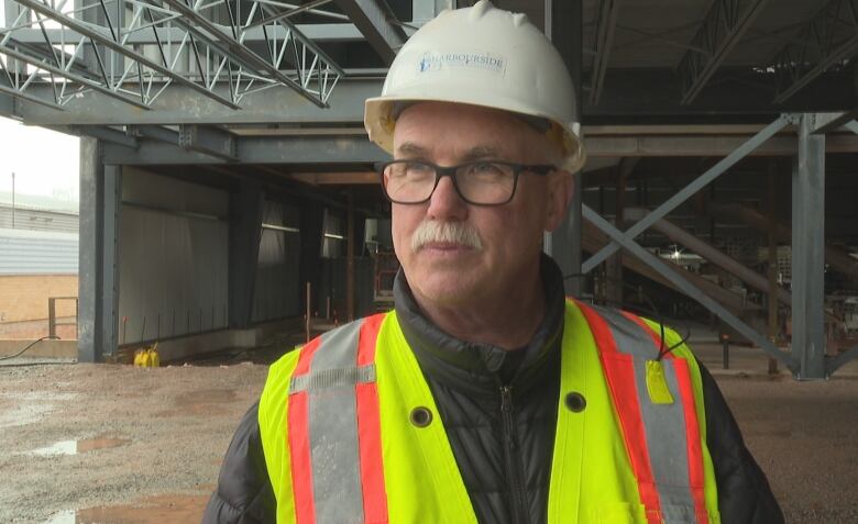 A person with a grey mustache wearing a white hard hat, and a bright yellow, orange and grey vest, stands in front of exposed beams at a construction site.