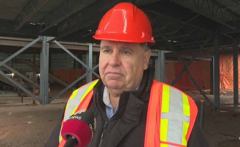 A person wearing an orange hard hat and a, orange, yellow, and grey vest, stands in front of exposed beams, and hanging orange tarps at a construction site.