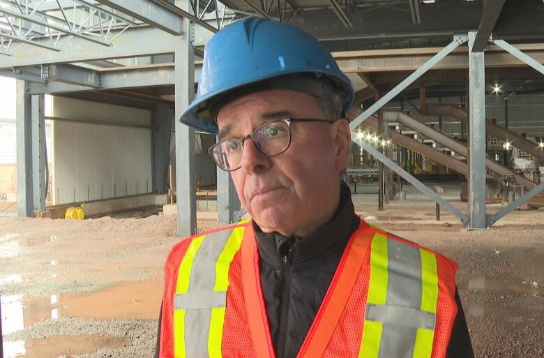 A man wearing eye glasses, a blue hard hat, and an orange, yellow and grey vest stands in front of exposed beams at a construction site.