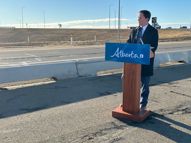 A man in a black coat standing at a podium on the side of a busy road with traffic. 