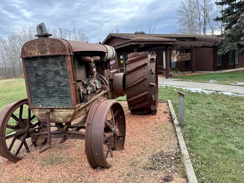 A large rusty tractor sits on the lawn near green grass under a blue sky next to a brown wooden museum. 