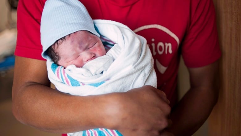 A man's arms are seen wrapped around a newborn baby swaddled in a white blanket with pink and blue stripes. 