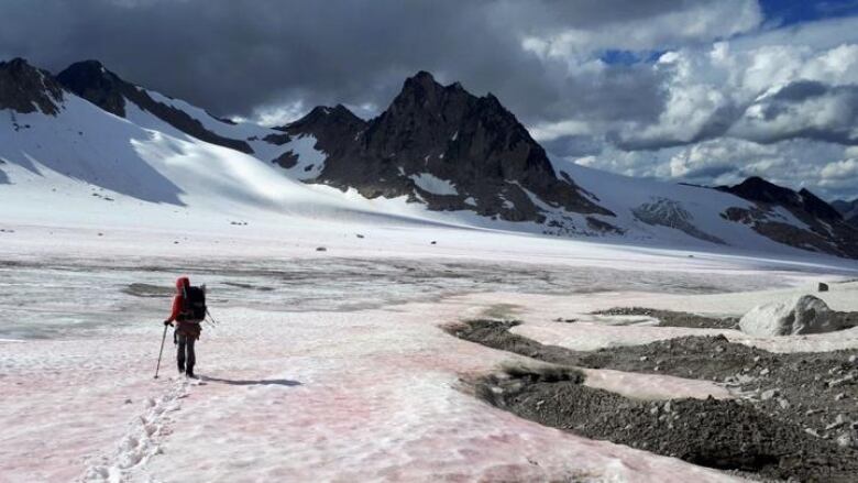 A hiker stands in a snowfield in front of a mountain range on a cloudy day