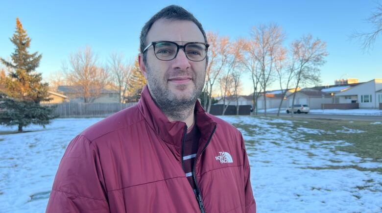 A man in a red jacket and glasses stands in a partly, snow-covered park in Regina. He's looking right to the camera around sunset.