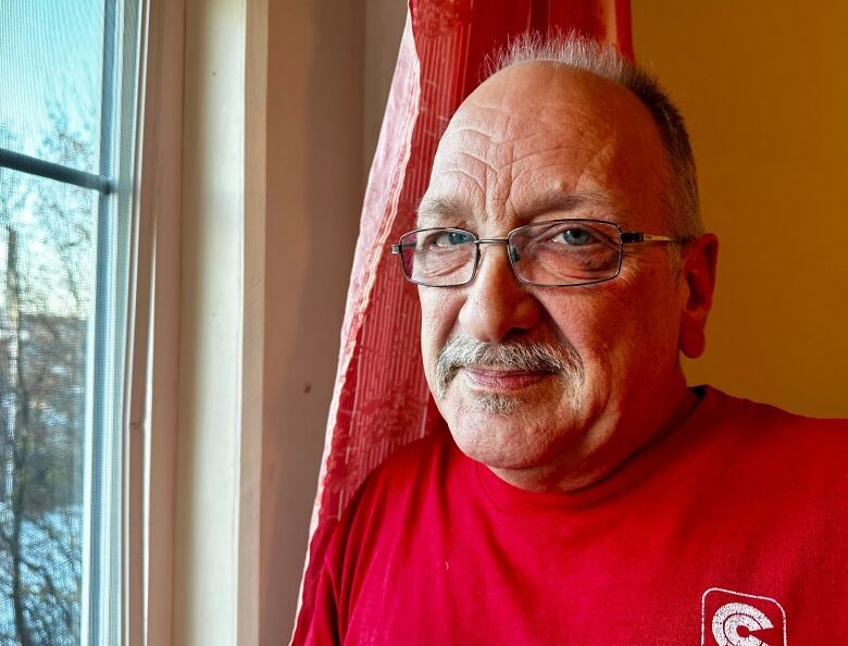 A man wearing glasses and a red shirt stands beside a window.