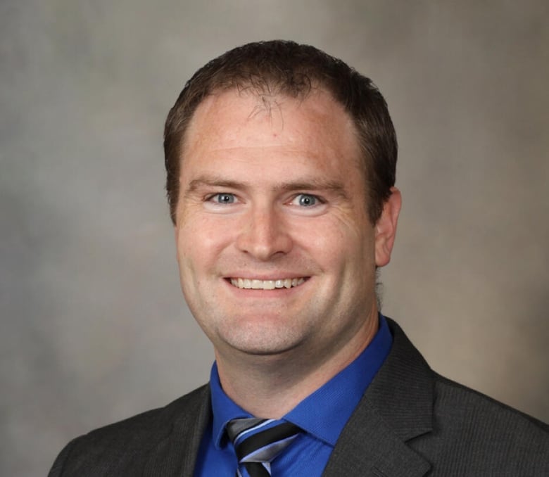 A man with blue eyes and short dark brown hair, wearing a dark grey blazer, smiles at the camera. He wears a royal blue button-up short with a diagonally striped tie.