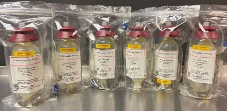 Several liquid medication bottles sit on a stainless steel countertop. They are inside sealed clear plastic bags and have red plastic caps. Their labels say Fluorouracil.