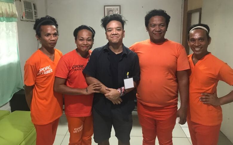 Four people wearing orange clothing stand on either side of a man wearing all black. All are smiling. 