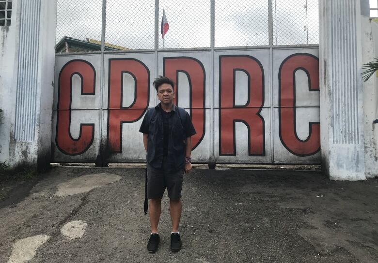 A man stands in front of a gate. On the gate are the letters CPDRC