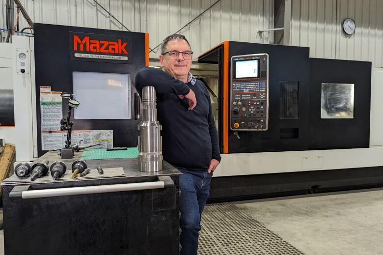 A man with short hair and glasses in a navy blue sweater and blue jeans stands in front of large, sophisticated piece of machinery in a shop. 