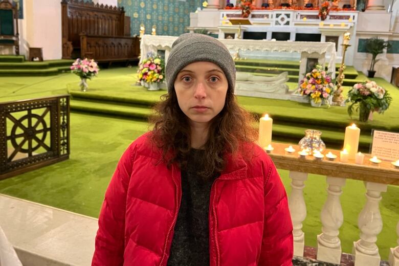 A woman in a winter coat and hat looks very serious in front of candles on an altar.