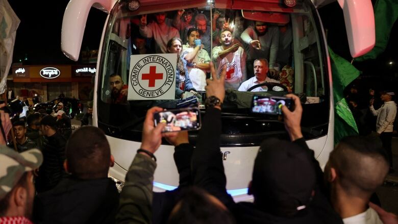 A crowded bus drives through the West Bank as hundreds of people surround it with green Palestinian flags.