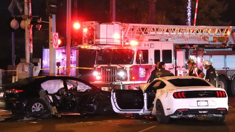 A black and a white car are pictured smashed and indented at an intersection with a fireftruck and emergency personnel behind them. 