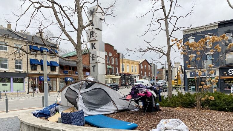 A tent and personal items like clothing, blankets, a lawn chair are seen in a raised garden bed with downtown businesses in the background