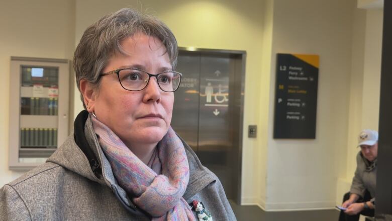 A woman with glasses, a scarf and a gray jacket stands near an elevator.