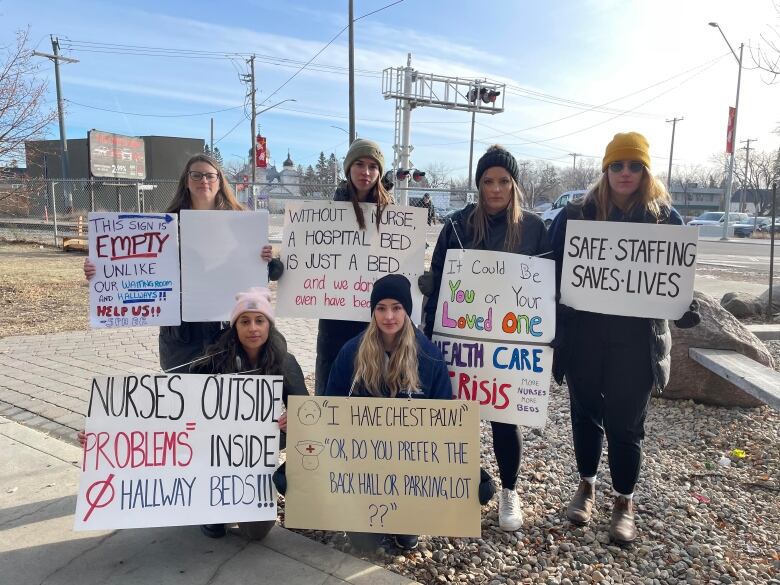 Four women stand with two women kneeling before them, all carrying signs that describe the situation in Saskatchewan hospitals, including a sign that says 