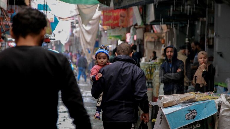 A man walks through a market in the rain with a little girl in his arms peering over his shoulder toward the camera. 