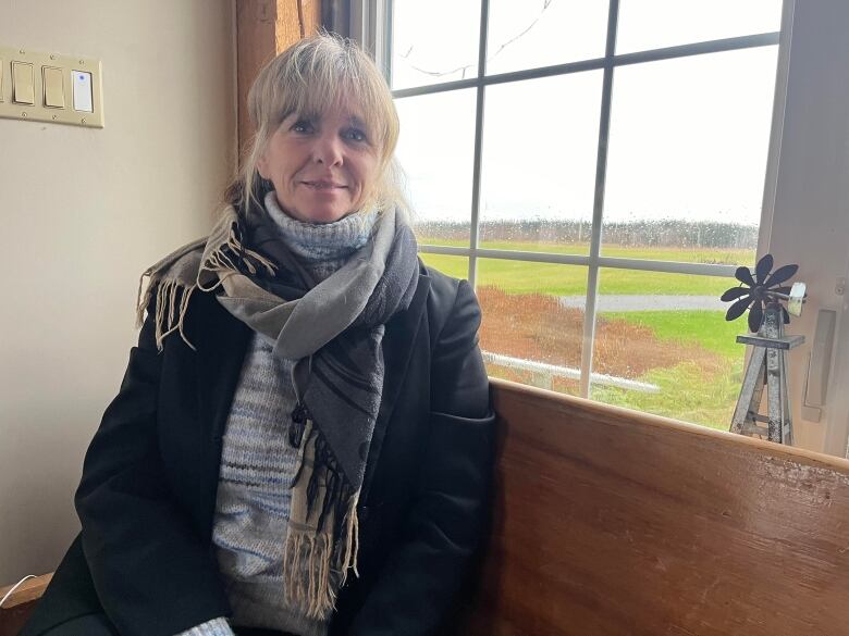 A middle aged woman with long blonde hair wears a scarf as she still infront of a window and smiles. 