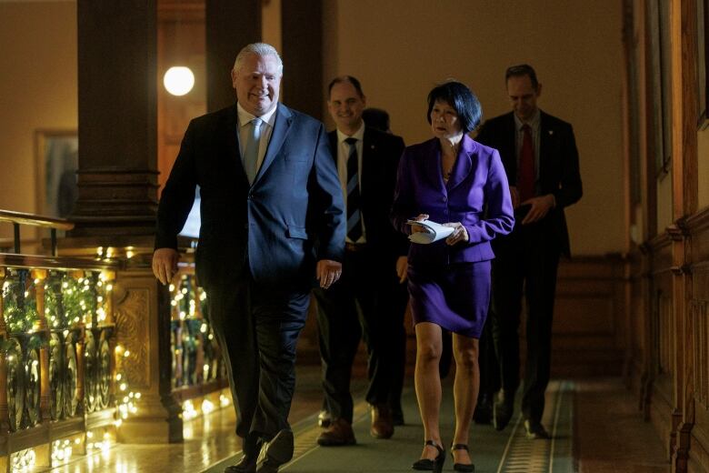 Doug Ford and Olivia Chow walk down an ornate, wood-paneled hallway with Christmas lights to their left. 