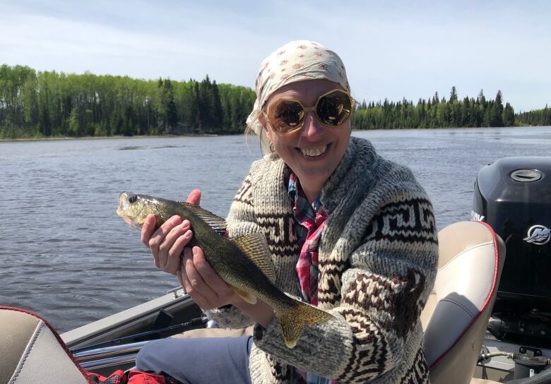 A woman wearing sunglasses, a head scarf and a Cowichan sweater sits in a boat on the water while smiling and holding a fish.