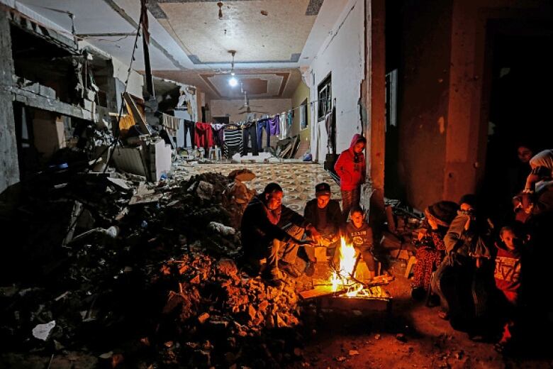 A group of people stand and sit around a fire. In the background, clothes hang from a line in a building that is nearly ruined. 