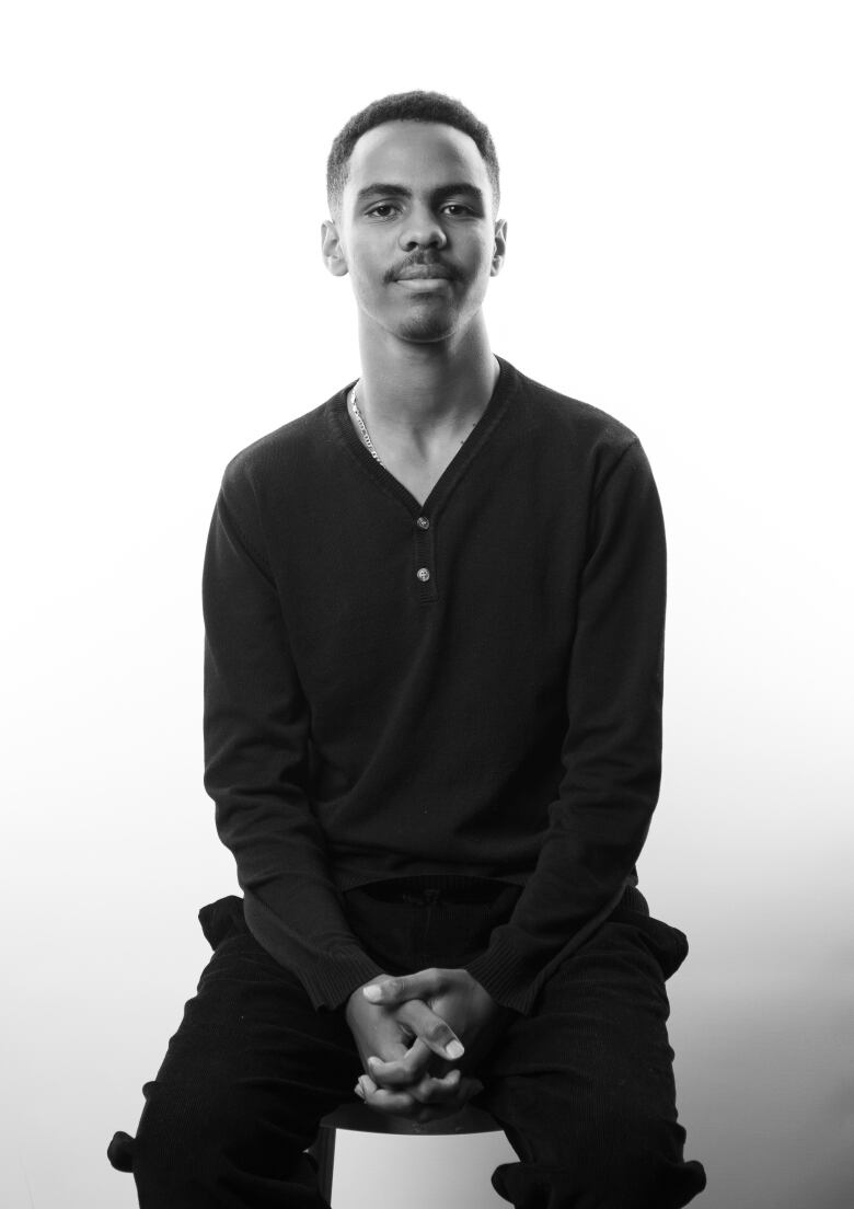 Black and white portrait of a man sitting on a stool with a closed mouth smile. He is clasping his hands on his lap. He is wearing a dark shirt with two buttons at the top; and dark pants.