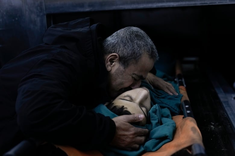 A man embraces and presses his face against the body of a deceased boy wrapped in a blanket on a stretcher.