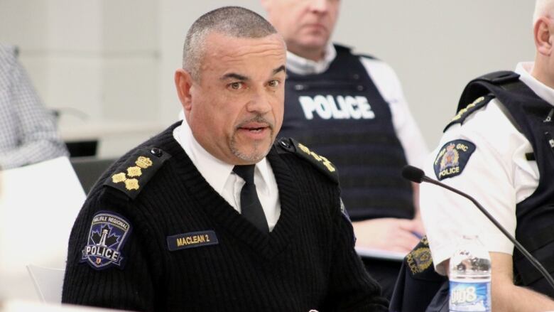 A Black man in a white collared shirt and tie under a black v-neck sweater with police badge on the arm sits at a table with people in chairs behind him