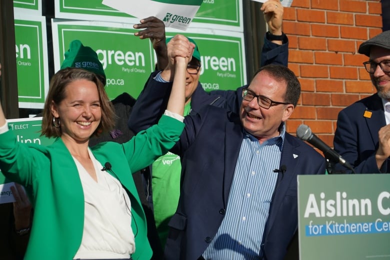 Woman and man hold hands up in a victory type pose at campaign stop with green signs in background