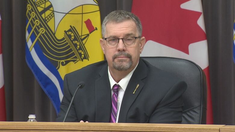 Man with short grey hair and glasses wearing navy suit jacket sits in front of New Brunswick flag and Canada flag.