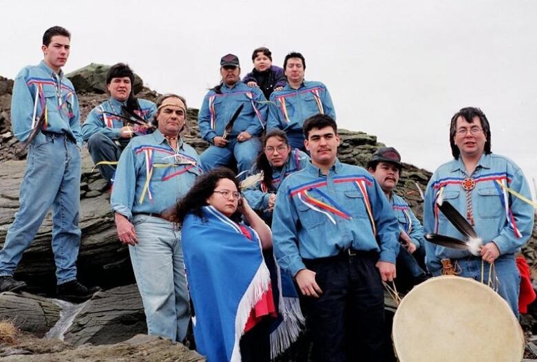 A group of men and women, dressed similarly in blue shirts and Mi'kmaw regalia and adornments, stand against a rocky outcrop.