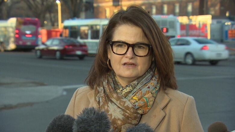 A woman in a coat stands outside on a busy street, speaking into reporters' microphones.