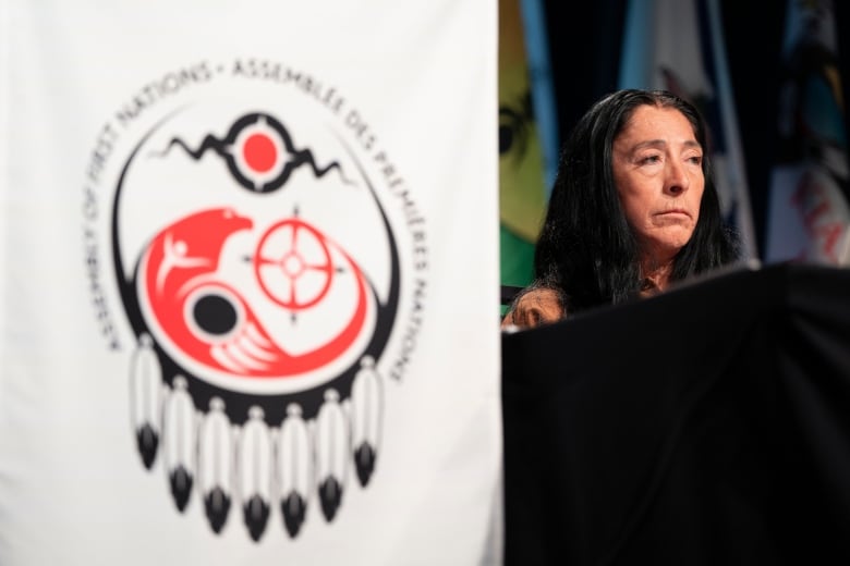 A woman sits on stage the background while the Assembly of First Nations logo is see out of focus in the foreground.