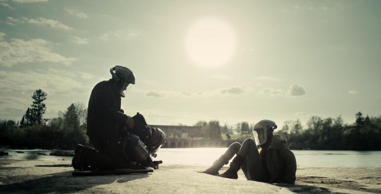 A still from a movie shows two people in helmets with face shields sitting in an open, snowy field, facing each other.