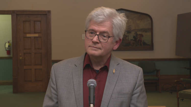 A man with white hair in a grey suit stands in front of a mic. 