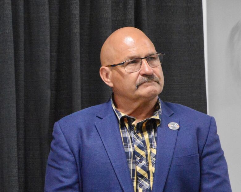 A man with a moustache wears a blue shirt with a yellow and blue plaid shirt underneath.