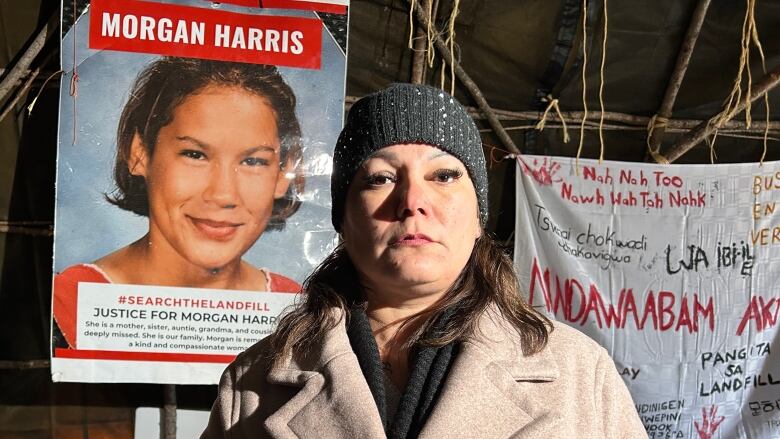 A woman wearing a toque and a cream-coloured wool coat is pictured inside a dark wigwam in front of a poster of a another woman which includes her name Morgan Harris.