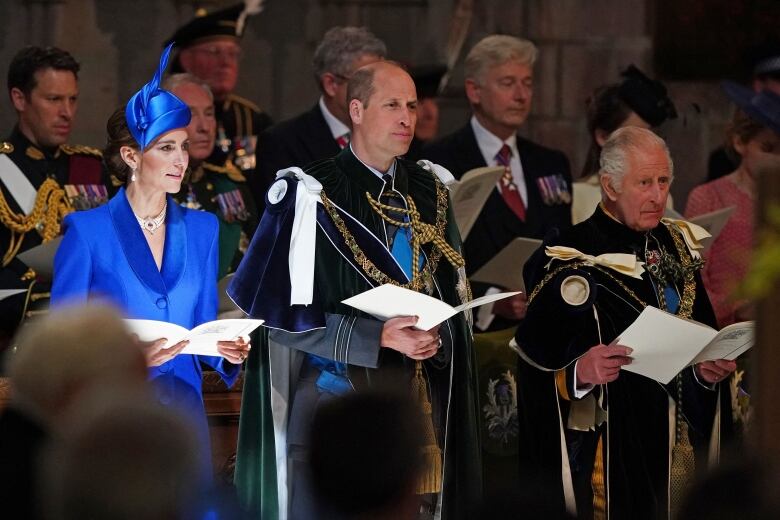 A woman in a blue outfit with a blue fascinator on her head stands next to two men in suits with robes tied around their shoulders.