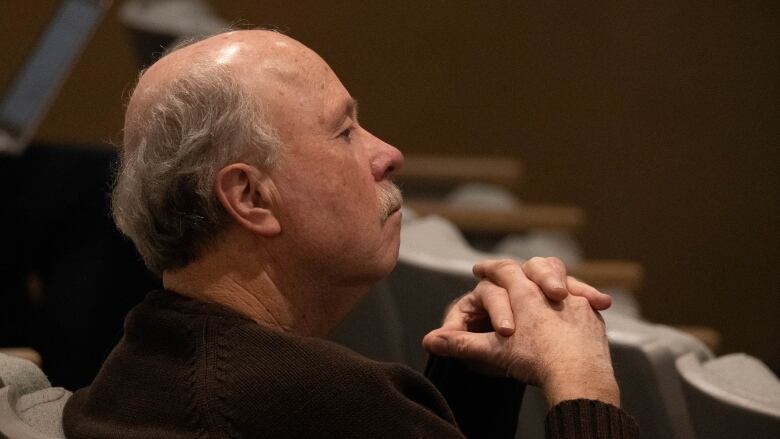 A man with white hair and a mustache watches from a seat in the audience with his hands tented