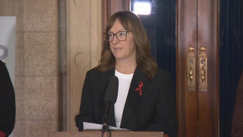 A woman with long hair, glasses and a black blazer speaks into a microphone