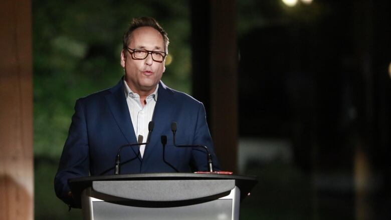 A man in a blue suit stands behind a podium and speaks.