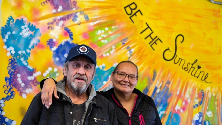 A man and woman stand in front of a bright wall that says be the sunshine.