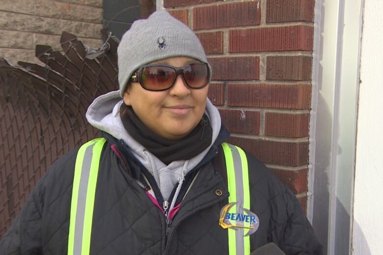 A woman wearing a safety jacket, toque and sunglasses stands on a sidewalk.