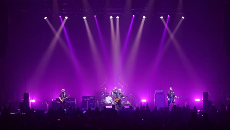 Three rock musicians play on stage inside a rink, with purple lighting in the background.