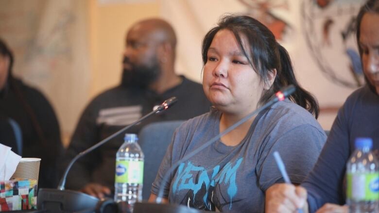 A woman in a t-shirt sits at a table with microphones. 