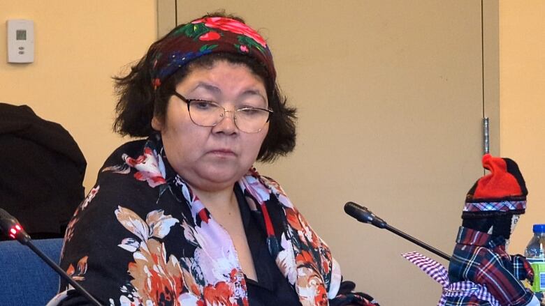 A woman wearing a scarf and floral top sits at a table with microphones. 