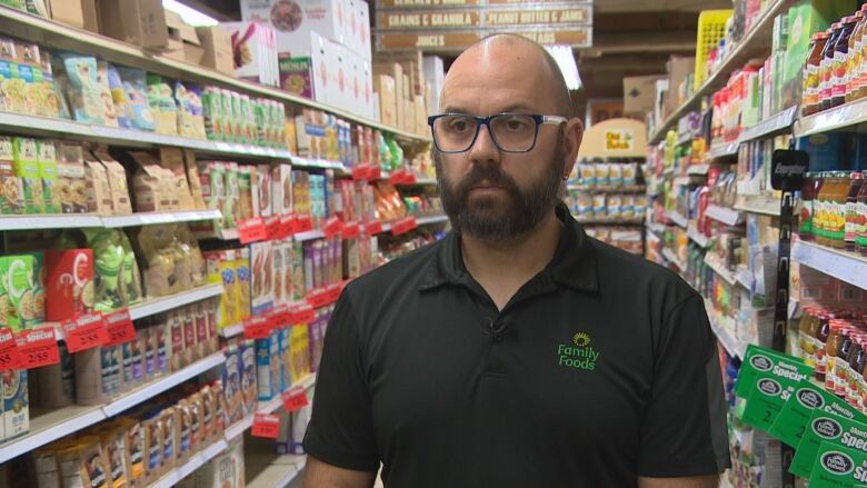 A man wearing a black polo shirt uniform with green embroidered letters standing in an aisle of a grocery store. 